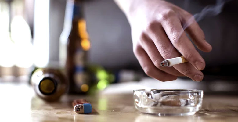 Close up a mano de varón sosteniendo cigarro encendido, al fondo botellas de cerveza.
